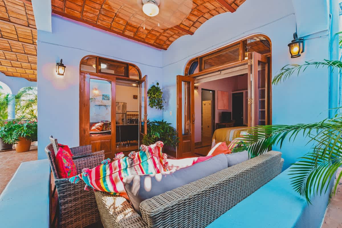 A cozy covered balcony with woven seating and colorful cushions opens directly into a bedroom through wide wooden doors. Potted palm plants add to the tropical ambiance, and the ceiling features exposed terracotta tiles above light blue walls.