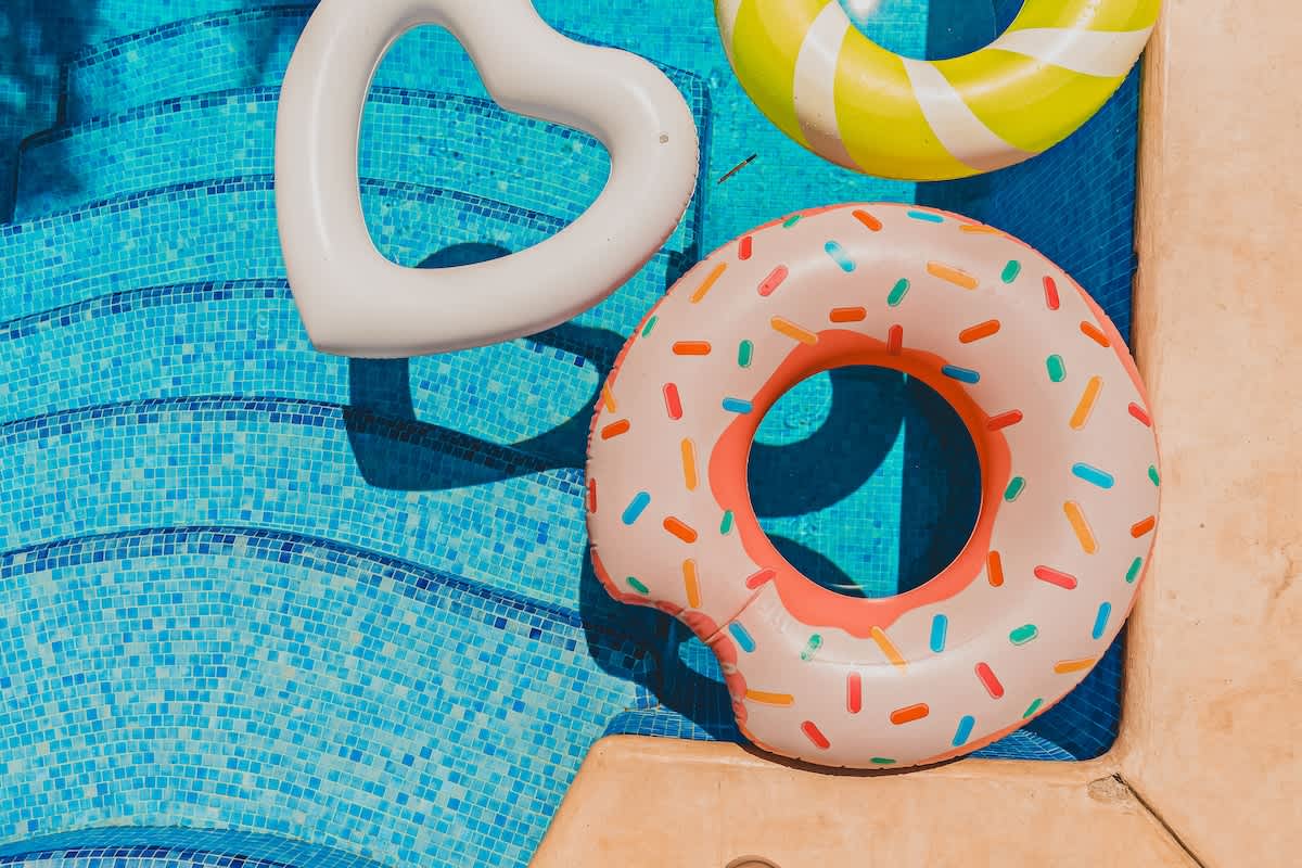 Two colorful inflatables, one shaped like a donut and another striped, float on the clear blue water of a swimming pool, with light blue mosaic tiles visible on the steps.