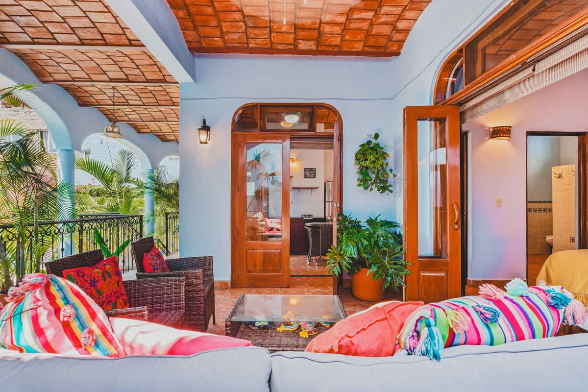 A vibrant covered balcony with a woven sofa and armchairs, adorned with bright pillows, faces a glass-top coffee table. Double wooden doors lead into an interior room, while lush greenery is visible beyond the railing, all under a terracotta-tiled ceiling.