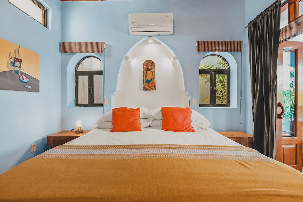 This vibrant bedroom showcases a large bed with orange pillows and a gold throw, set against light blue walls with an arched white headboard featuring a colorful artwork. Two arched windows flank the bed.