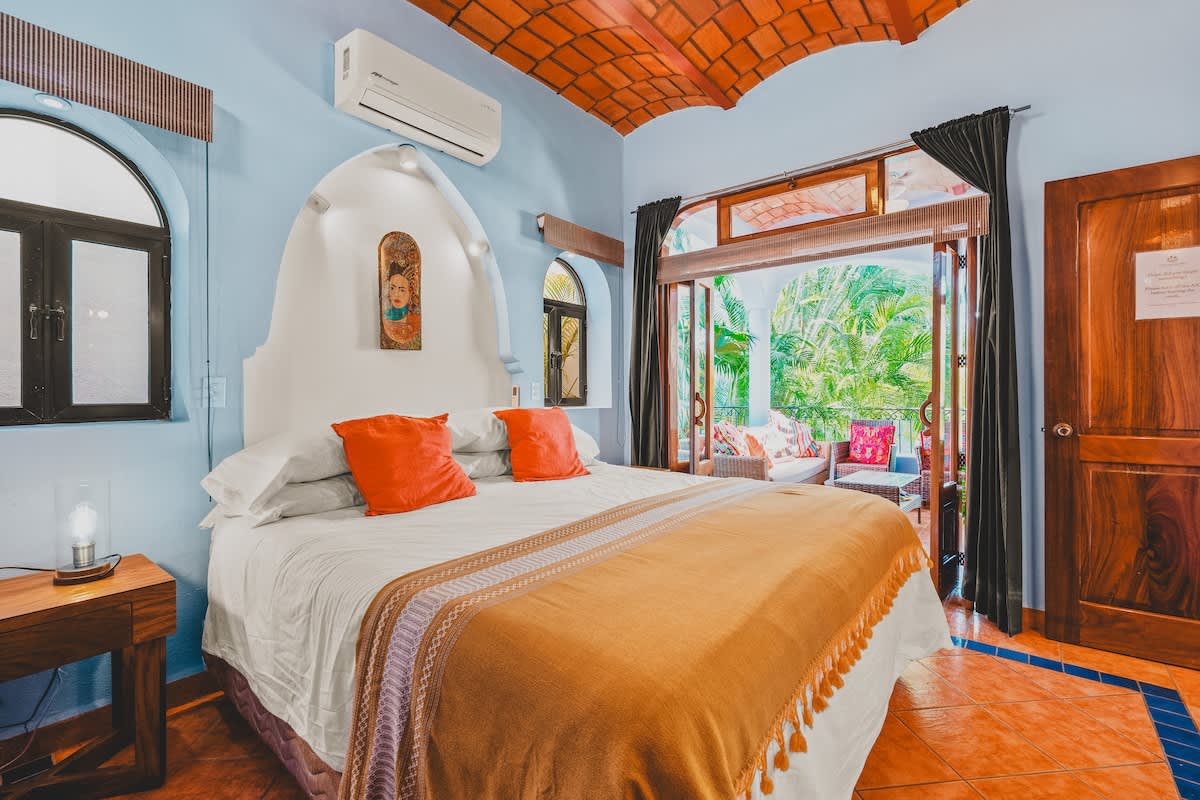 A colorful bedroom with light blue walls and a terracotta vaulted ceiling features a large bed with orange pillows and a gold throw, flanked by arched windows. A door opens to a lush outdoor balcony.