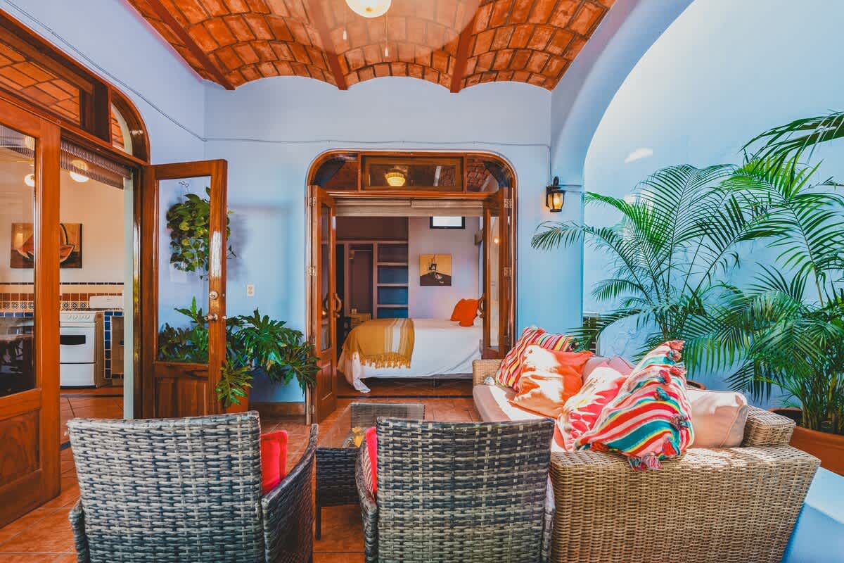 A covered outdoor balcony with woven chairs and a sofa, decorated with bright pillows, looks into a bedroom with a white bed and orange accents through an open wooden doorway. Lush green plants are placed around the patio, complementing the terracotta-tiled ceiling.