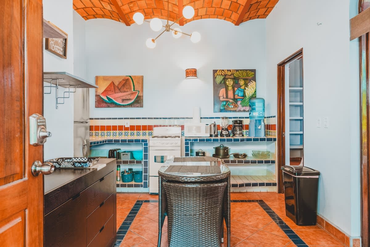 A bright and colorful kitchen boasts tiled countertops with blue and red accents, a white stove, and a stainless steel refrigerator. A wicker dining table is centrally placed, illuminated by a modern chandelier and flanked by vibrant artwork.