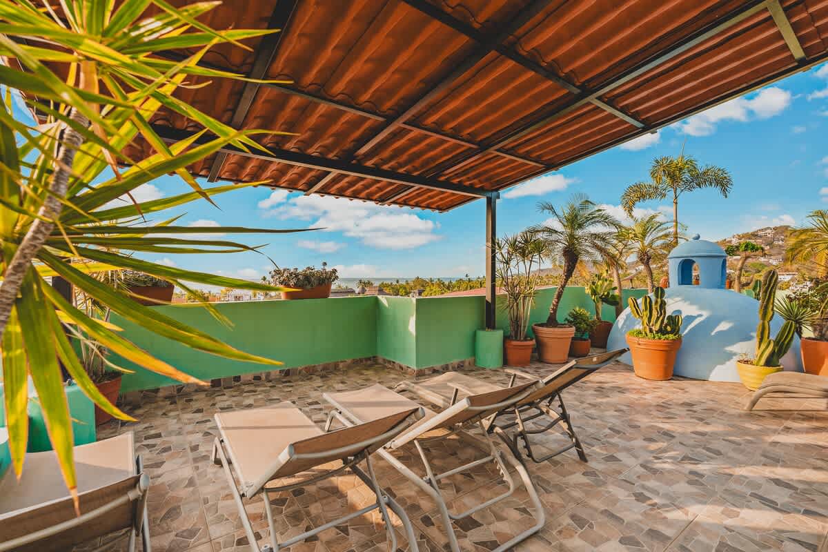 A sunny rooftop deck features terracotta-tiled flooring, several lounge chairs, and potted plants including cacti and palms. A corrugated metal roof provides partial shade, offering views of the ocean and distant hills under a bright blue sky.