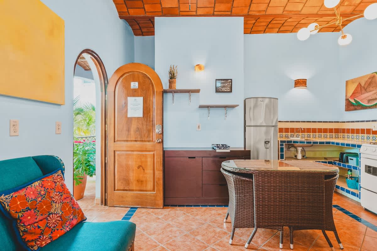 An open-concept living and dining area features light blue walls, terracotta tile flooring with blue accents, and a vaulted ceiling with exposed brick. A teal sofa with a colorful throw pillow sits near a wooden arched door, while a wicker dining table and chairs are positioned next to the kitchen entrance.