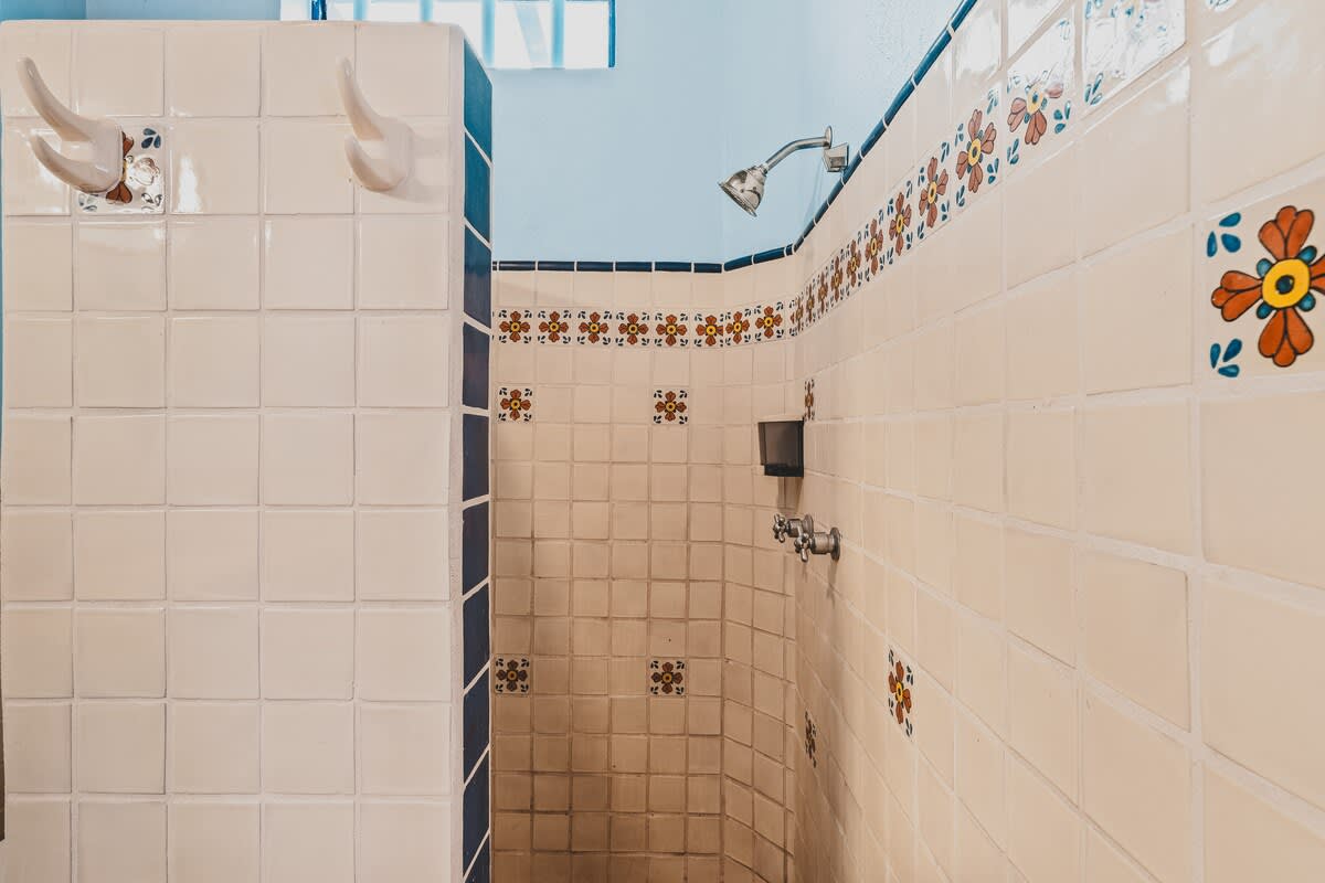 A brightly tiled shower stall features white square tiles with decorative floral accents and a blue border, complementing the light blue walls. A chrome shower head is mounted on the wall, and white hooks are visible on the adjacent tiled wall.