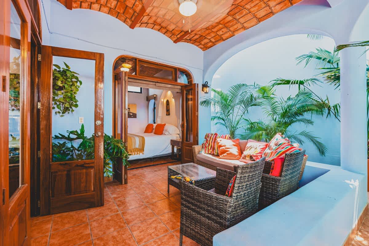 A charming covered balcony with woven seating and vibrant cushions offers a direct view into a bedroom featuring a white bed with orange throw pillows. The balcony is adorned with potted plants and has a terracotta-tiled ceiling, creating an inviting space.
