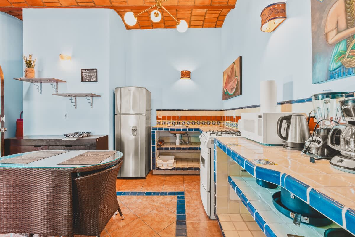 A vibrant kitchen features light blue walls, terracotta tile flooring with blue accents, and a vaulted ceiling with exposed brick. The space includes a stainless steel refrigerator, a white stove, and a counter with blue and white tiled backsplash, equipped with various small appliances.