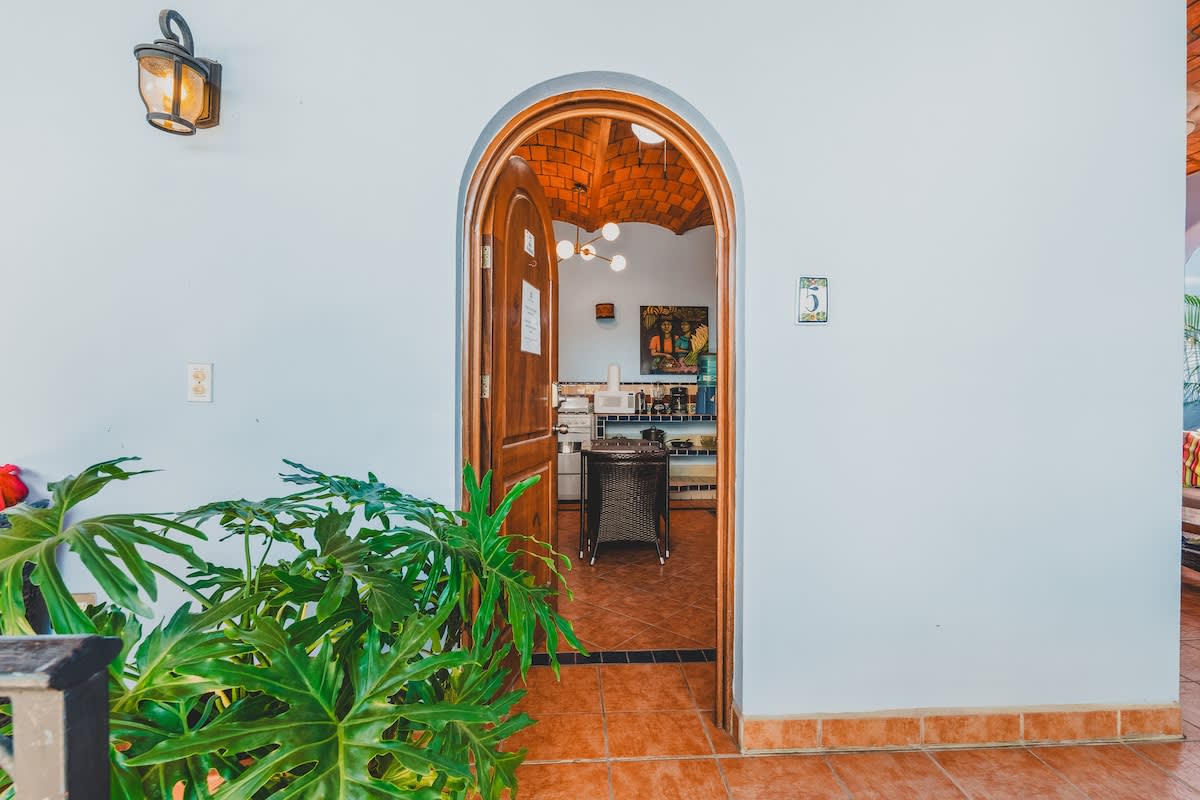 A light blue wall with an arched wooden doorway leads into a brightly lit interior space with terracotta tile flooring and a view of a kitchen or dining area. Lush green plants frame the entrance, and a rustic wall sconce provides exterior lighting.