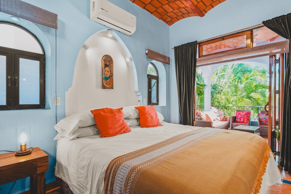 A bright bedroom with light blue walls and a terracotta vaulted ceiling features a large bed with orange accents and direct access to a lush outdoor balcony with seating. An air conditioning unit is mounted above the bed.