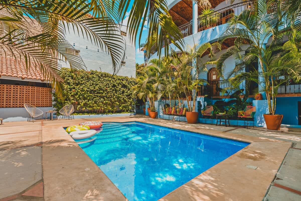 A rectangular swimming pool with inflatables is the focal point of a sunny courtyard, surrounded by a concrete deck, lush green walls, and potted palm trees, with a light blue building in the background.