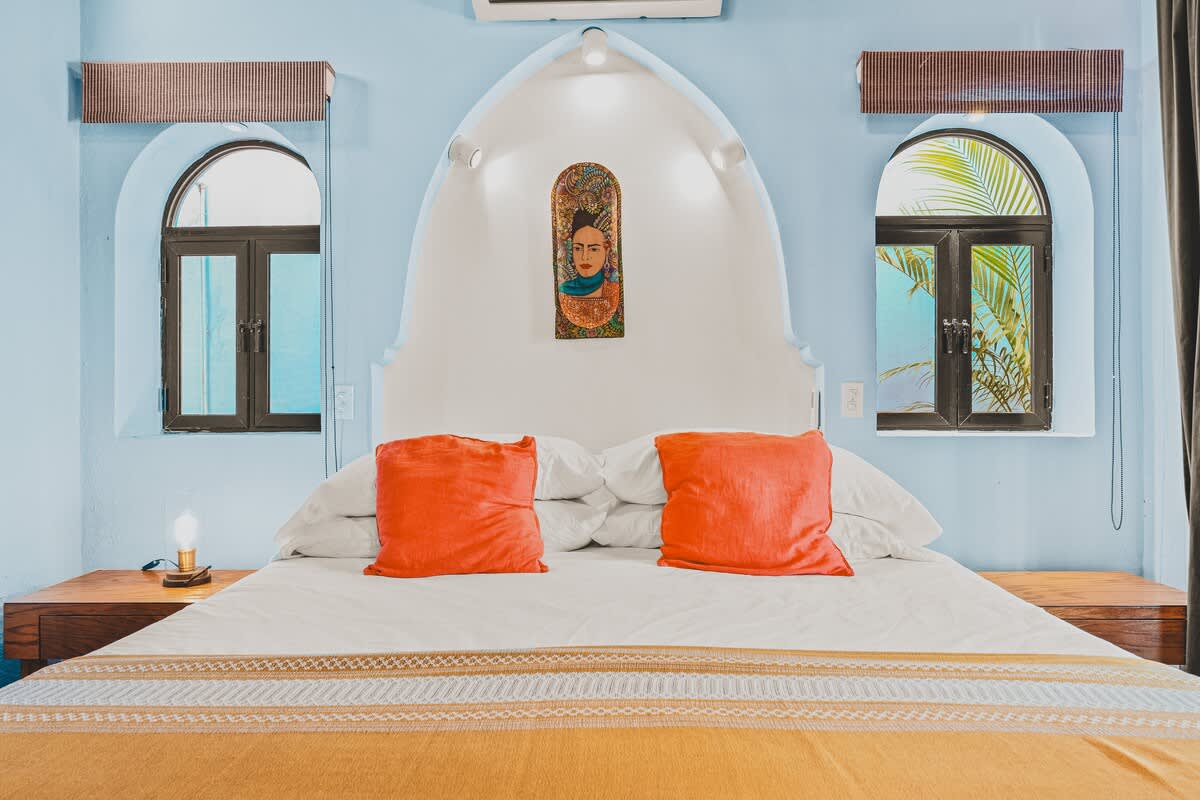 A cozy bedroom features a large bed with white linens, orange pillows, and a gold patterned throw, centered against a unique arched white headboard with a decorative art piece. Light blue walls surround two arched windows.