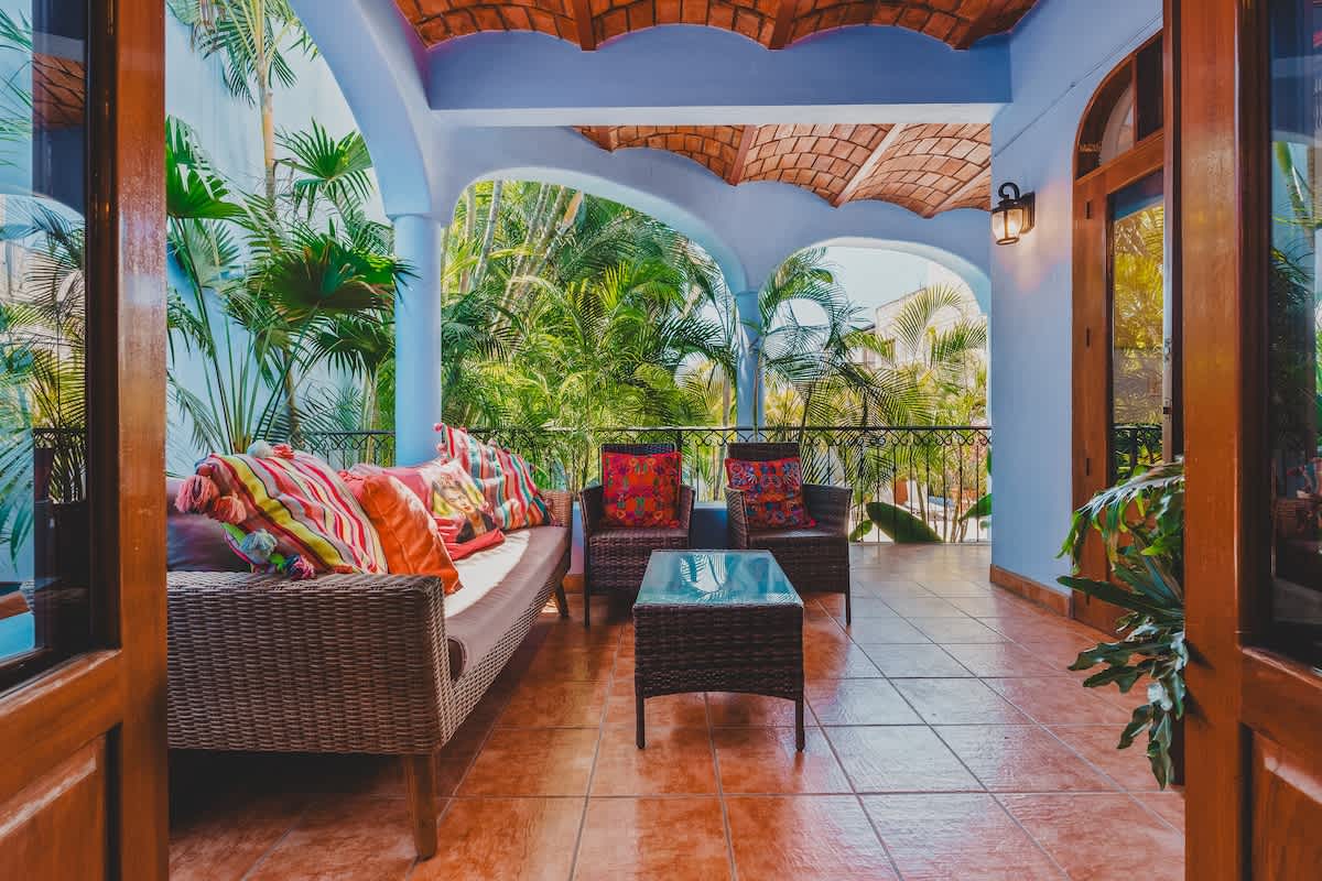 A spacious covered balcony features a woven sofa and armchairs with colorful pillows, centered around a glass-top coffee table. A wrought-iron railing overlooks lush tropical foliage, while the ceiling boasts exposed terracotta tiles and light blue walls.