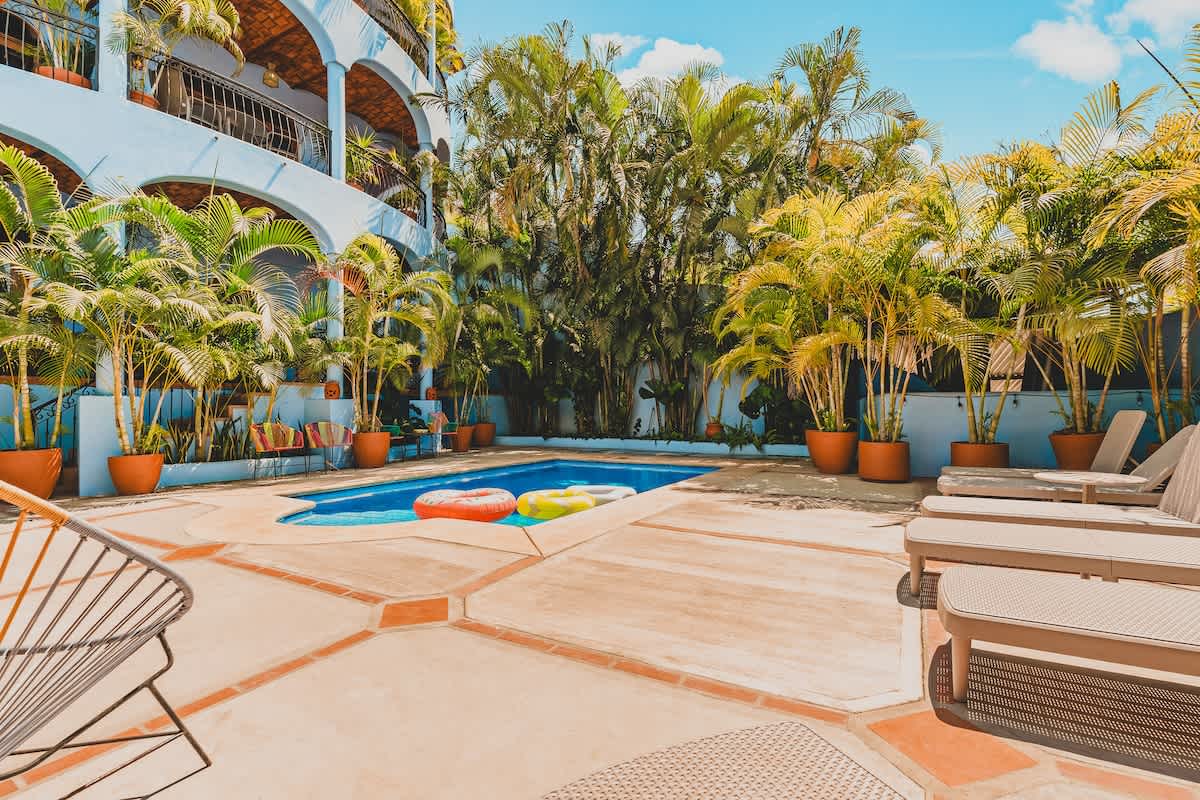 A rectangular swimming pool with blue water is centered in a paved courtyard, surrounded by potted palm trees and lounge chairs. A light blue building with arched balconies is visible on the left.