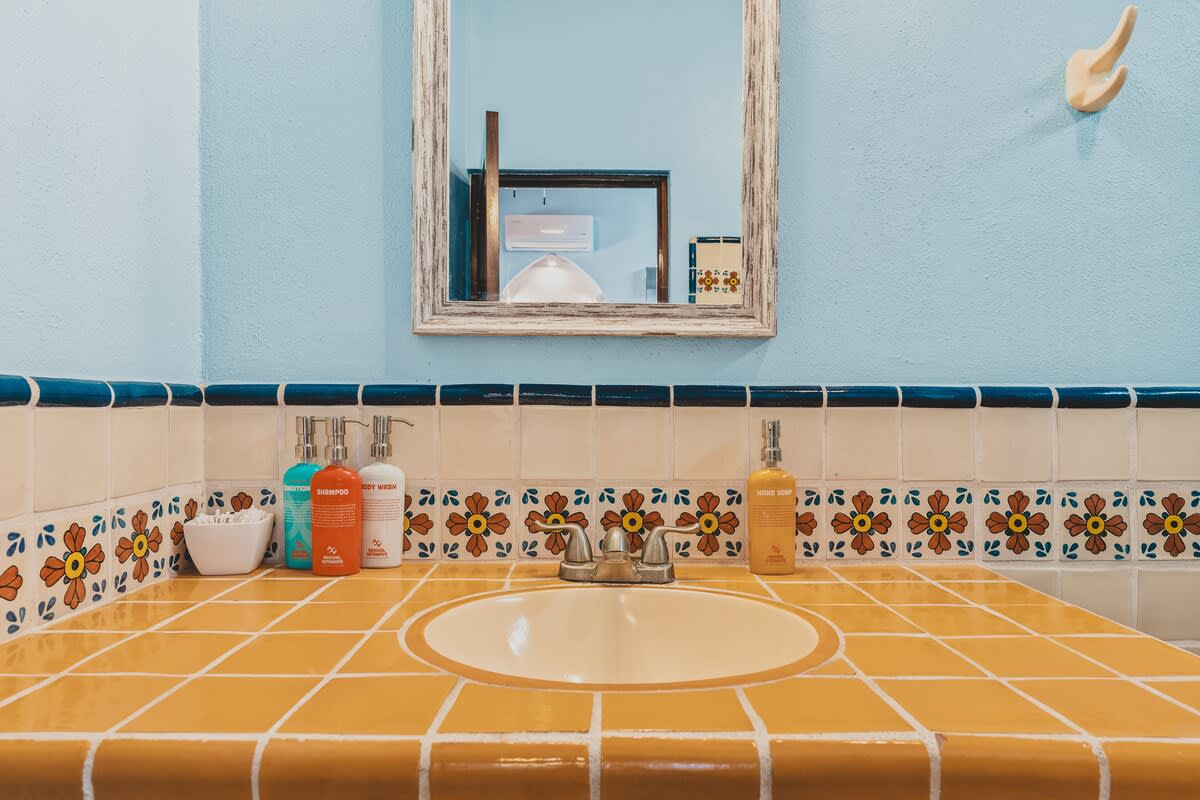 A vibrant bathroom vanity features a yellow tiled countertop with a white oval sink, surrounded by decorative floral tiles and a blue border. A rustic wooden mirror hangs above, reflecting the light blue wall, with various toiletries neatly arranged.