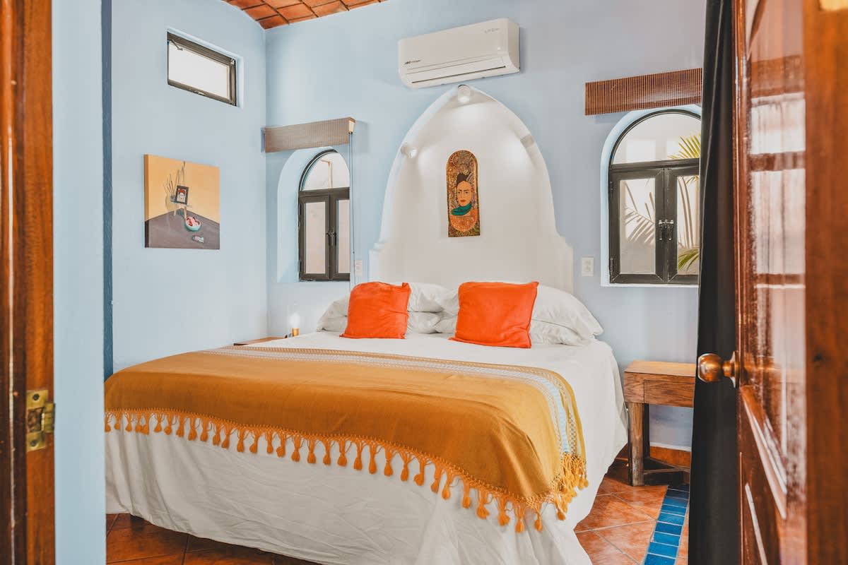 A welcoming view into the bedroom reveals a large bed with white linens and orange pillows, flanked by arched windows. Light blue walls and a unique arched headboard create a vibrant atmosphere.