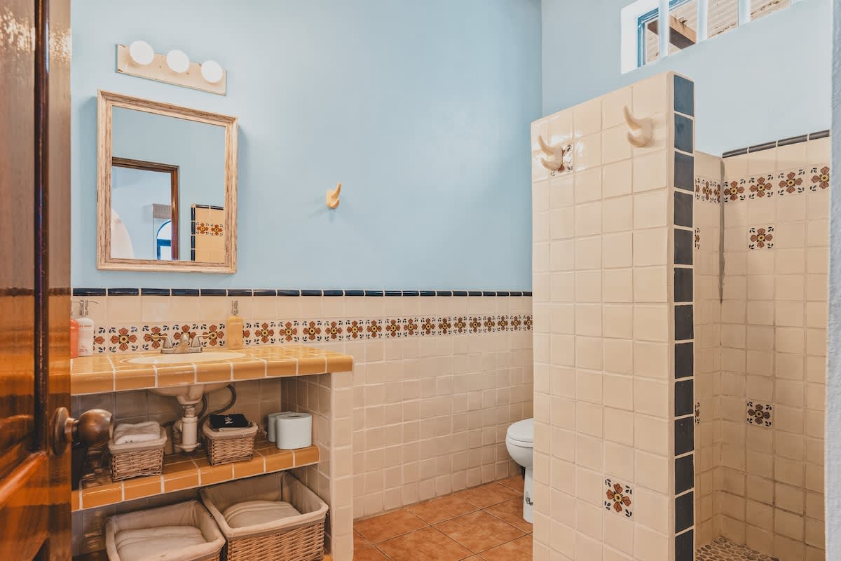 A vibrant bathroom features light blue walls, a tiled vanity with a wooden mirror, and a tiled shower stall with decorative accents. Woven baskets are stored under the sink.
