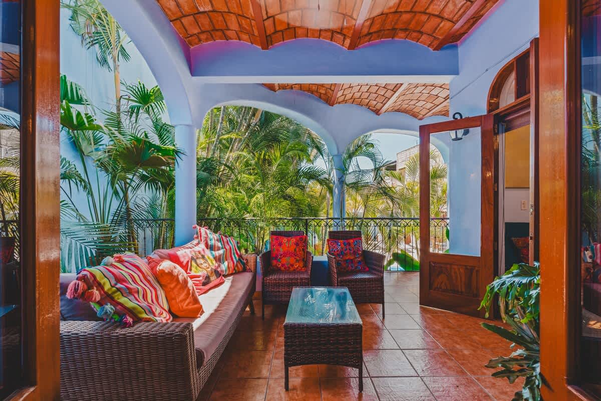 A spacious covered balcony with light blue walls and a terracotta-tiled ceiling features a woven sofa and armchairs with bright pillows. It overlooks a lush green landscape through a decorative metal railing, offering an inviting outdoor retreat.