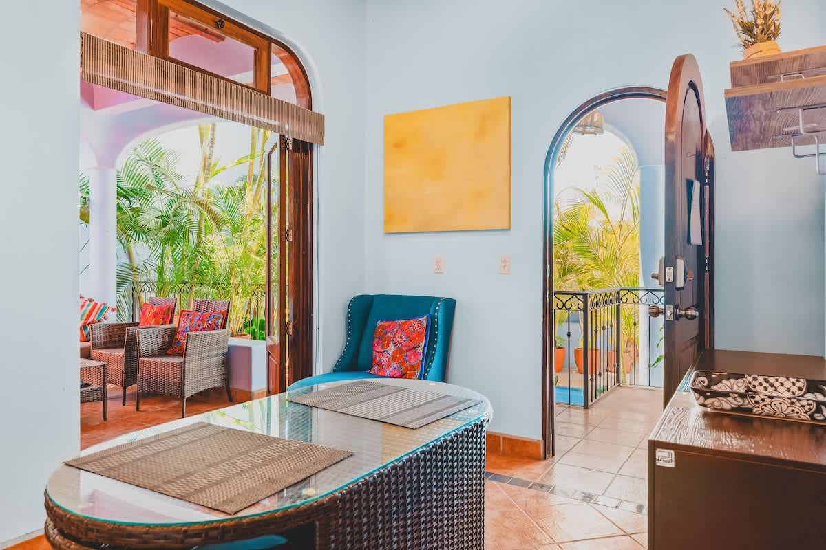 A dining area features a wicker table with a glass top and placemats, adjacent to a blue armchair. An arched doorway leads to a lush outdoor balcony with additional seating.