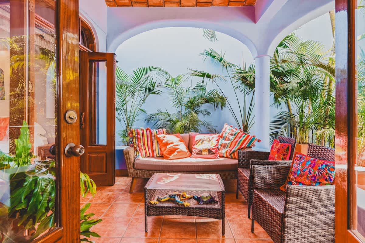 A covered outdoor balcony with a woven sofa and armchairs, featuring colorful striped pillows, is surrounded by lush tropical plants. The balcony has a terracotta-tiled ceiling and light blue walls, creating a serene outdoor living space.