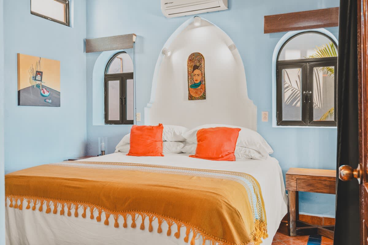 This inviting bedroom features a large bed with white bedding and bright orange pillows, centered against an arched white headboard with a decorative art piece. Two arched windows and light blue walls complete the vibrant space.