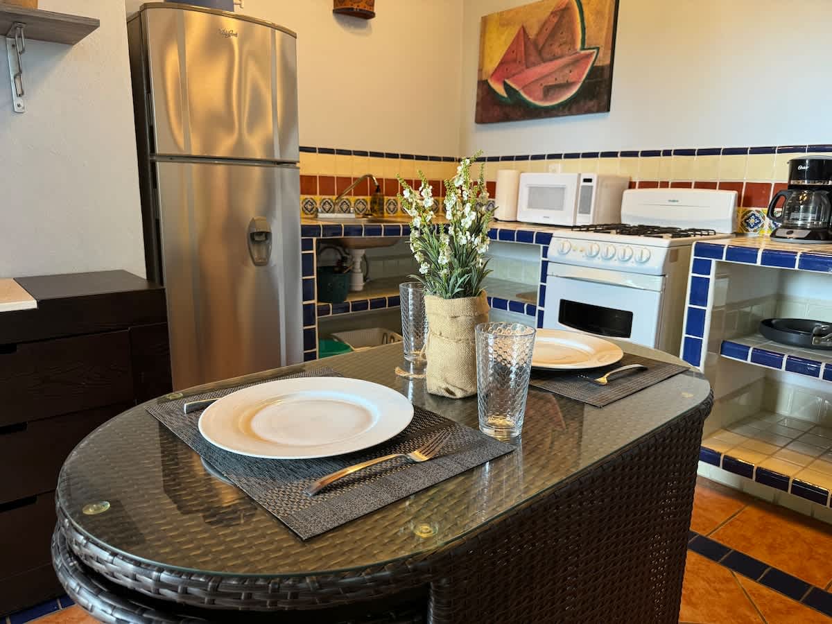 A small dining area features a glass-top table with two place settings, adjacent to a kitchen with a stainless steel refrigerator, microwave, and stove, all with colorful tiled accents.