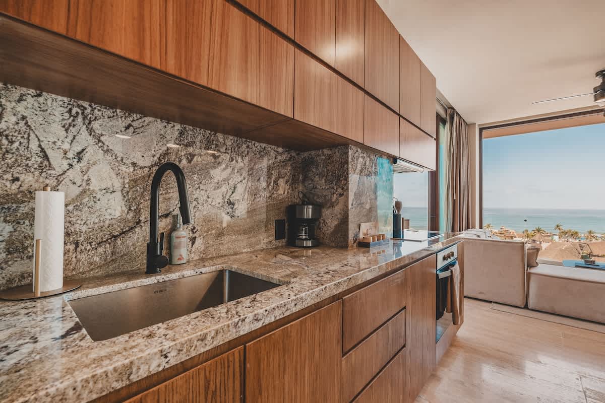 Spacious kitchen with light granite countertops and wooden cabinetry, featuring a stainless steel sink, a black coffee maker, and an open layout connecting to a living area with ocean views.