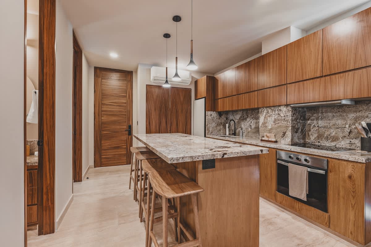 Sleek kitchen with light wood cabinetry, a large granite island with wooden bar stools, and integrated stainless steel appliances including an oven and cooktop.