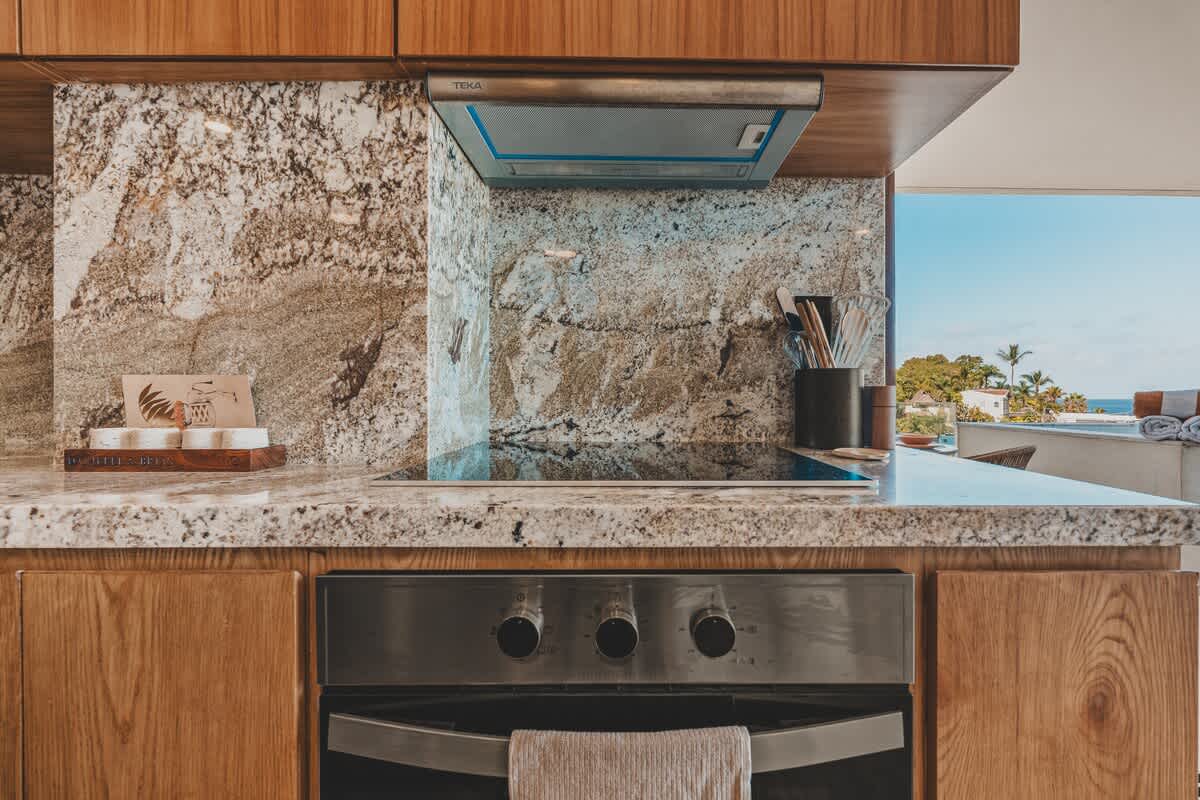 Modern kitchen with light granite countertops and backsplash, featuring a built-in electric stovetop, an oven below, and a stainless steel range hood, with wooden upper and lower cabinets.