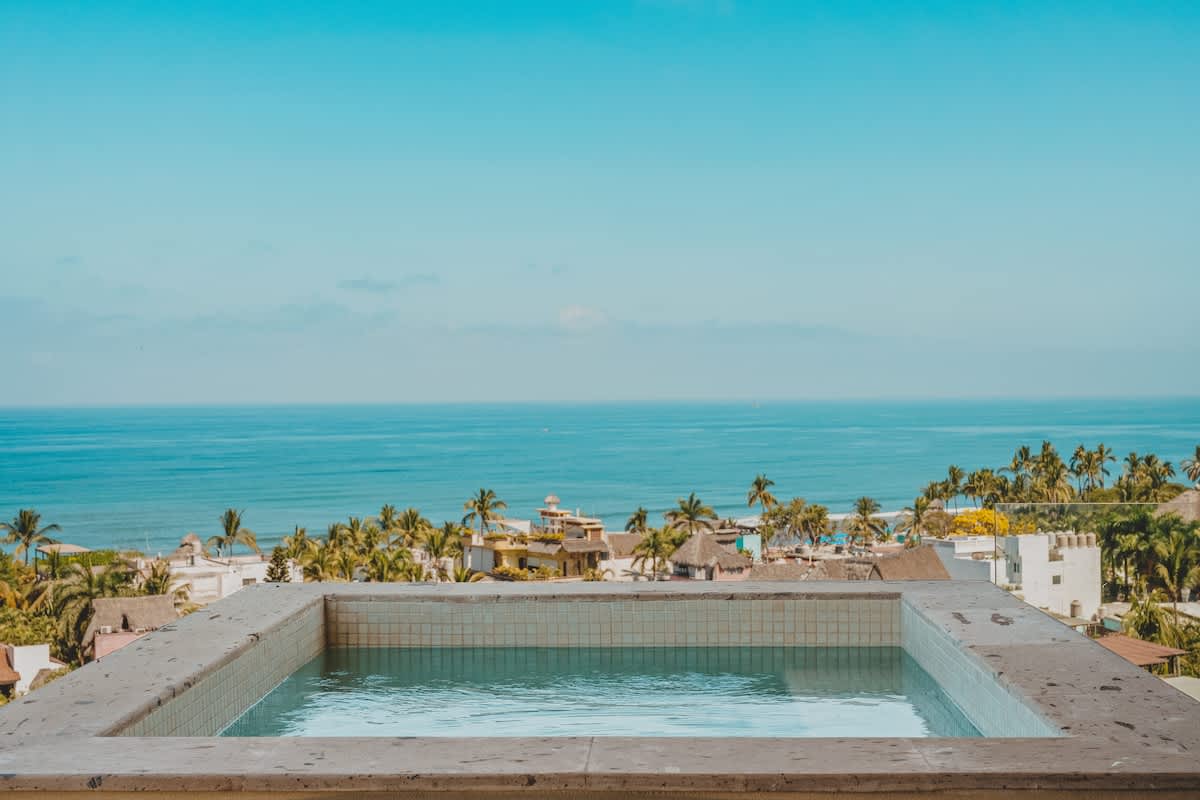 A square, tiled hot tub is positioned on an elevated terrace, offering an unobstructed, expansive view of the calm blue ocean and a lush coastal town dotted with palm trees.