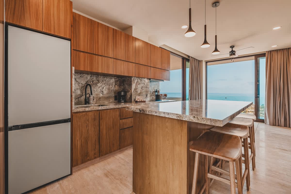 Modern kitchen with light wood cabinetry, a large stainless steel refrigerator, granite countertops, and a sink, illuminated by pendant lights.