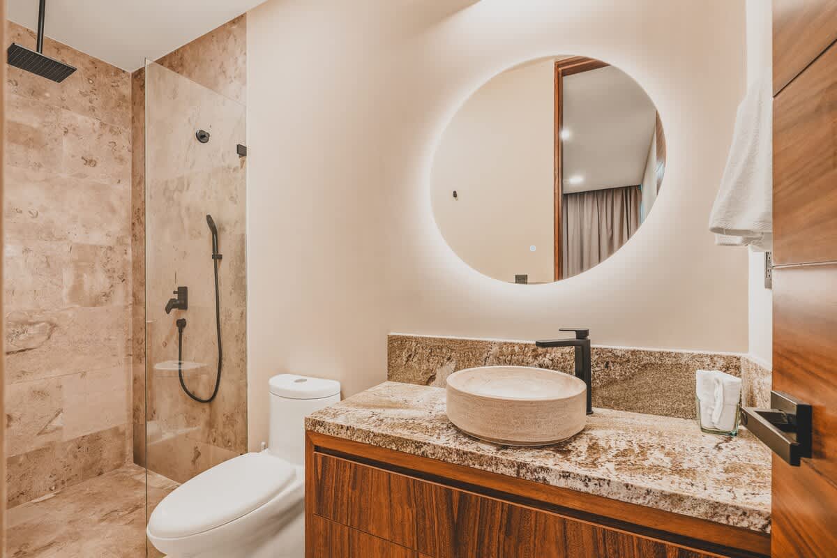 A modern bathroom featuring a walk-in shower with light marble-patterned tiles and black fixtures, alongside a floating wooden vanity with a stone countertop and a round vessel sink. A large, backlit circular mirror adds a contemporary touch.