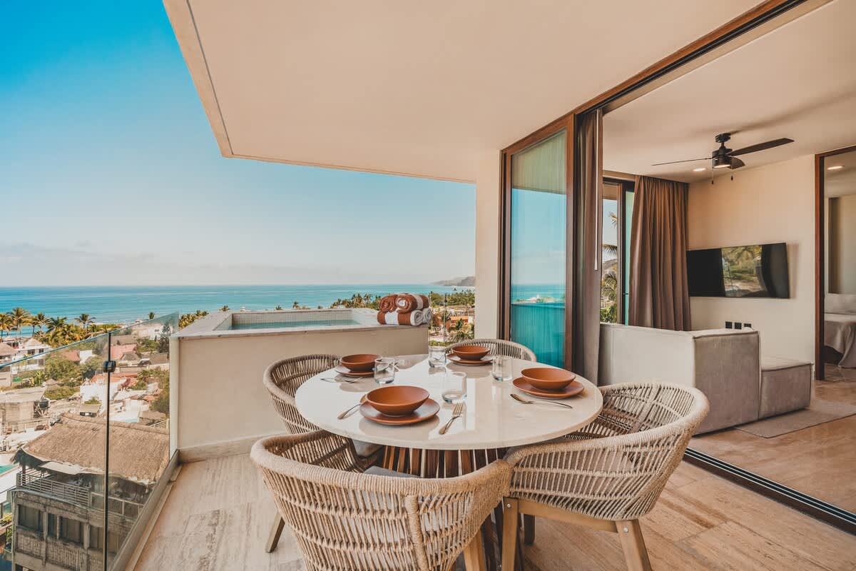 A private balcony offers an outdoor dining area with a round table and woven chairs, alongside a small hot tub, all with a breathtaking view of the ocean and the coastal town below.