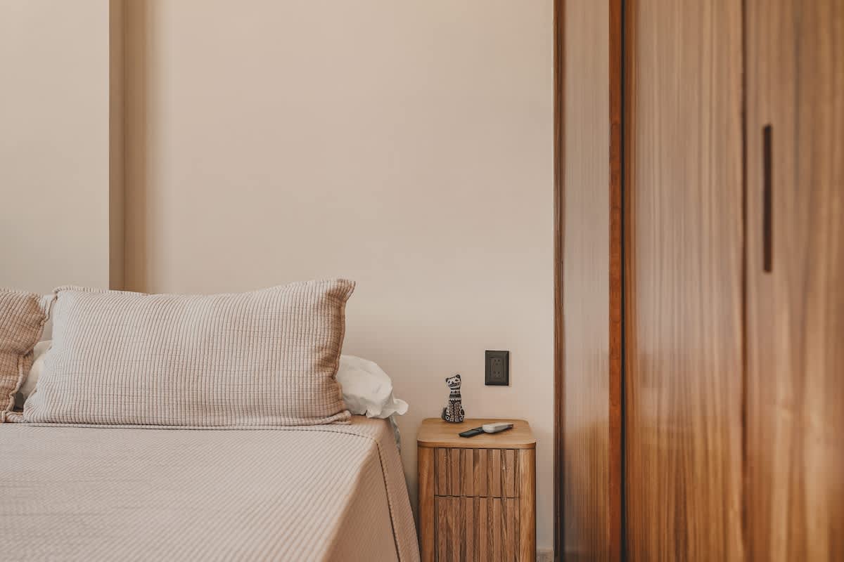 Bedroom corner with light beige walls and bedding, featuring a wooden nightstand with a small decorative sculpture and a knife, next to a built-in wooden wardrobe.
