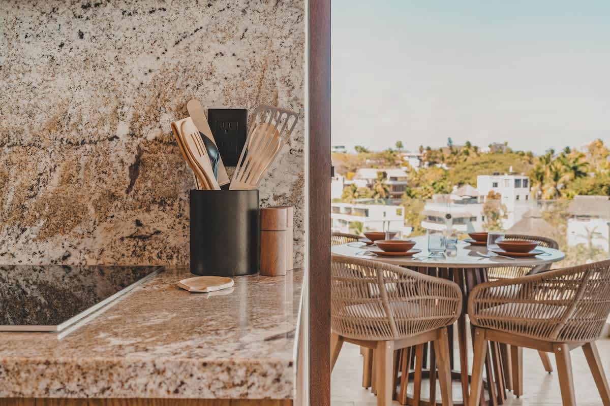 Kitchen counter with light granite, showing a black utensil holder with wooden tools, next to a glass sliding door leading to an outdoor dining area with wicker chairs and a view of the city and ocean.