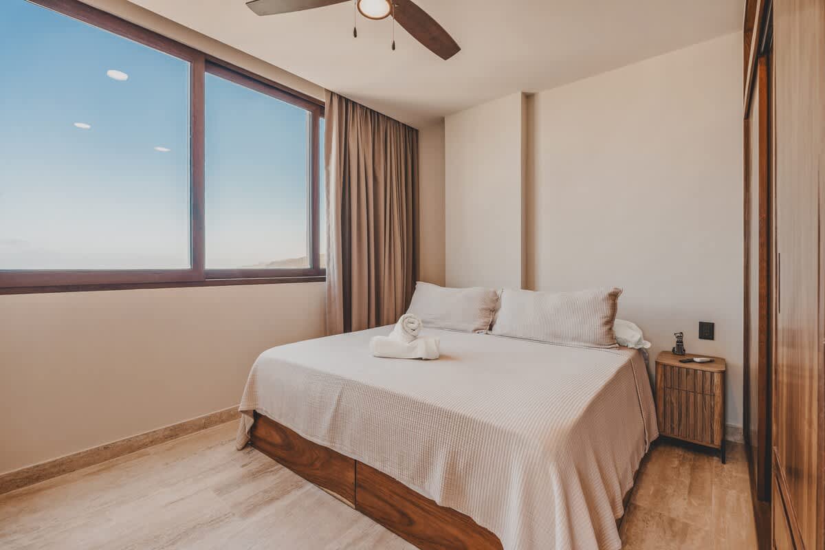 Bedroom with a queen-sized bed, light bedding, and two rolled white towels, featuring a large window with light brown curtains, a wooden nightstand, and a ceiling fan, offering a bright and airy feel.