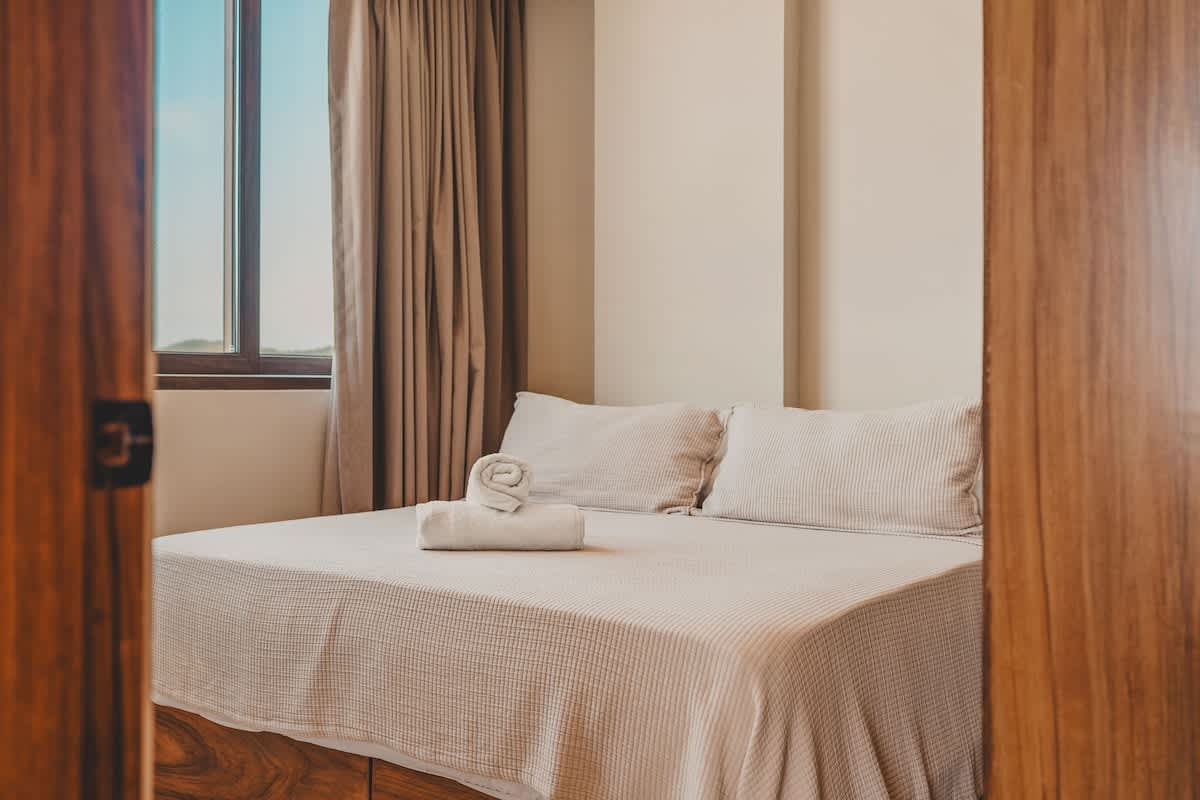 A cozy bedroom featuring a queen-sized bed with a light-colored textured bedspread and two pillows, accented by a rolled white towel. A large window with brown frames and light beige curtains provides natural light, complementing the neutral wall tones.