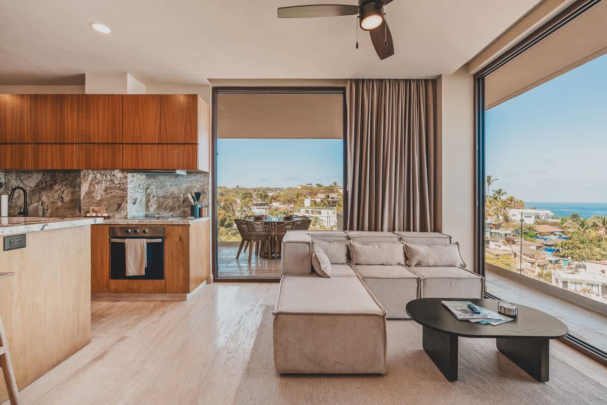 Open-concept living area with a light-colored sectional sofa, a dark coffee table, and large sliding glass doors leading to a balcony with outdoor seating and sweeping views of the ocean and surrounding town.