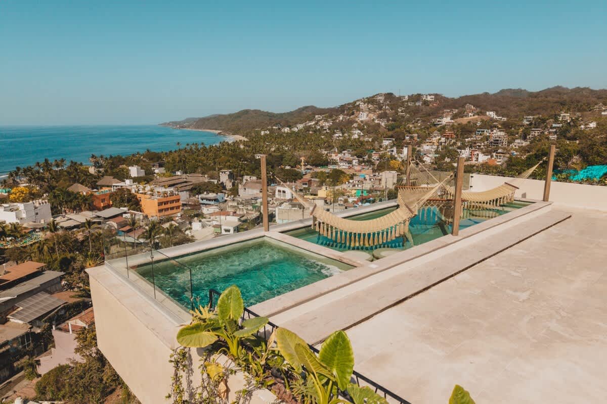 An elevated rooftop pool area featuring two small pools with woven hammocks suspended over the water, offering panoramic views of a vibrant coastal town, lush hills, and the expansive blue ocean under a clear sky. Green plants adorn the concrete deck.