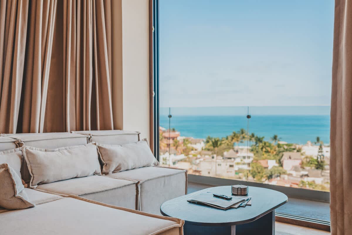 Bright living room with a light-colored sectional sofa and a dark oval coffee table, offering panoramic ocean and city views through a large floor-to-ceiling window with beige curtains.