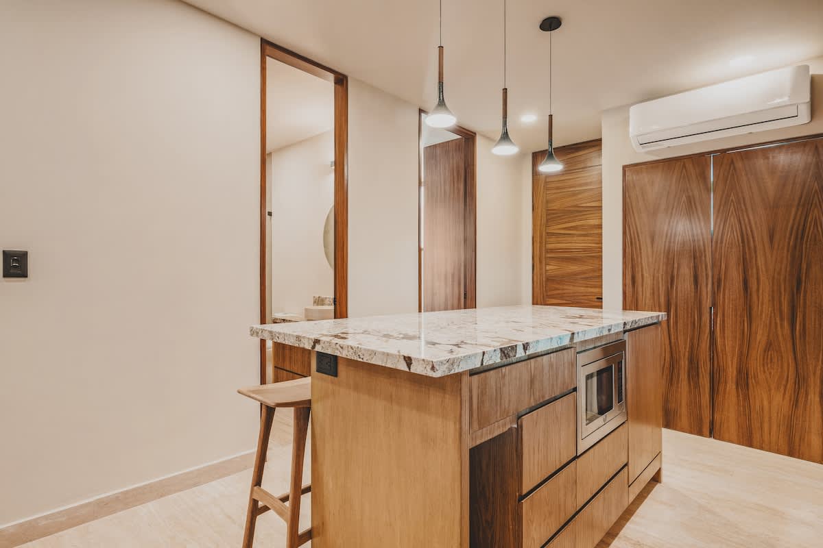 Contemporary kitchen island with a light granite countertop, integrated microwave, and wooden cabinetry, illuminated by three pendant lights. A single wooden bar stool is visible.