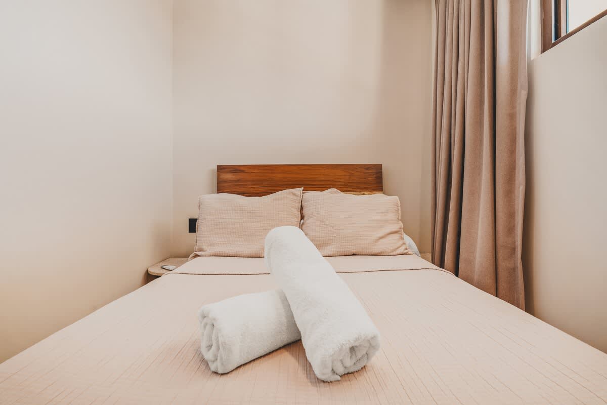 Serene bedroom with a wooden headboard and light beige bedding, accented by two neatly rolled white towels, with a light-colored wall and a glimpse of beige curtains.