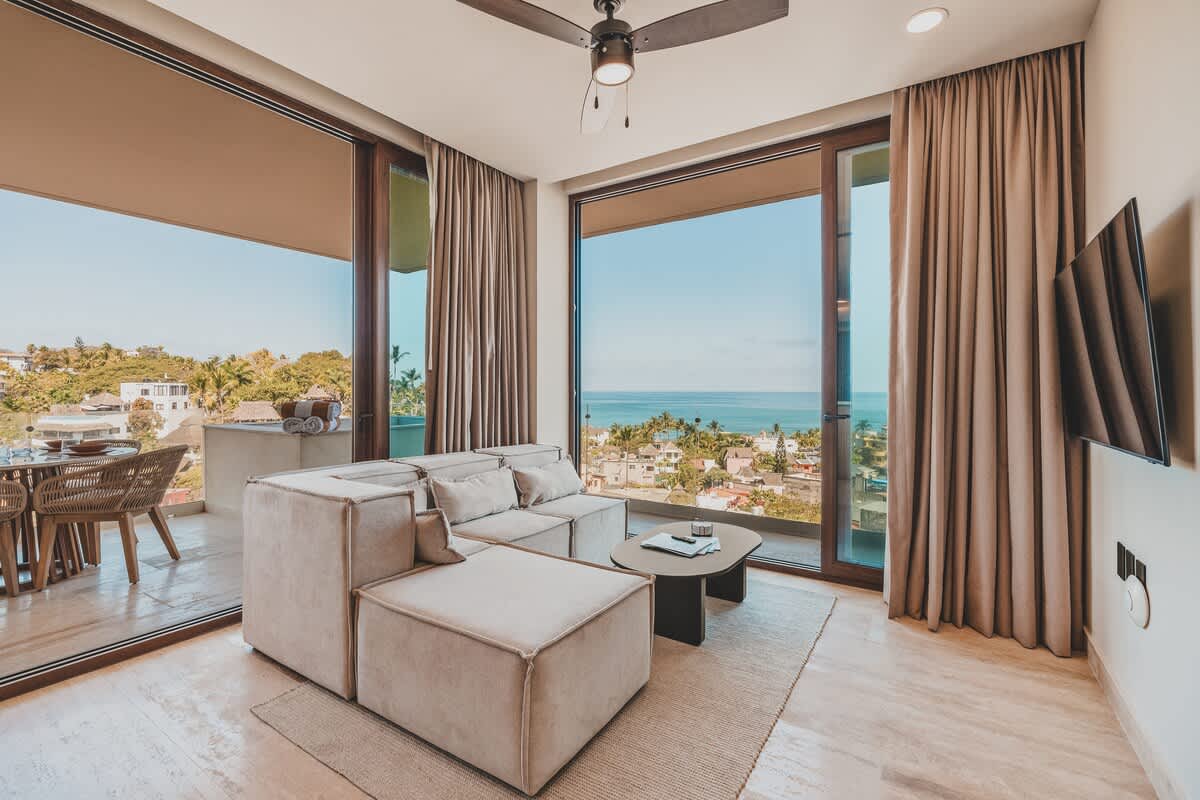Expansive living room with a light beige sectional sofa and a dark coffee table, featuring a ceiling fan and large sliding glass doors opening to a balcony with sweeping ocean and city views.