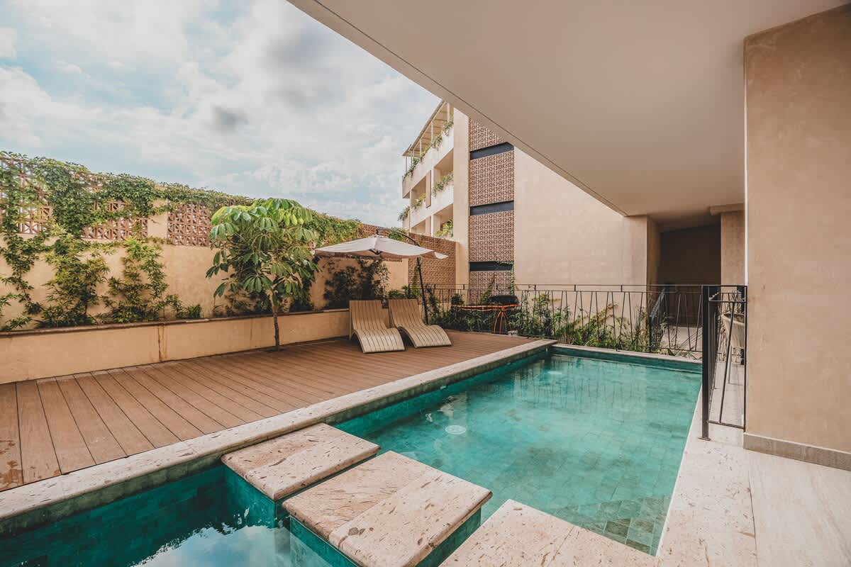 Expansive swimming pool area with a wooden deck, two lounge chairs under an umbrella, and stone steps leading into the turquoise water, bordered by a textured wall with climbing vines.
