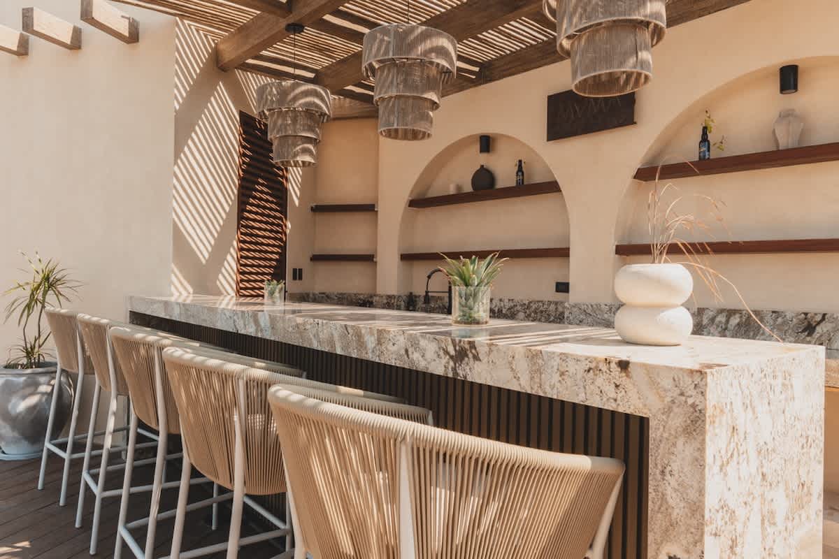 A stylish outdoor bar features a speckled marble countertop with several woven bar stools, illuminated by unique hanging light fixtures under a slatted wooden pergola.
