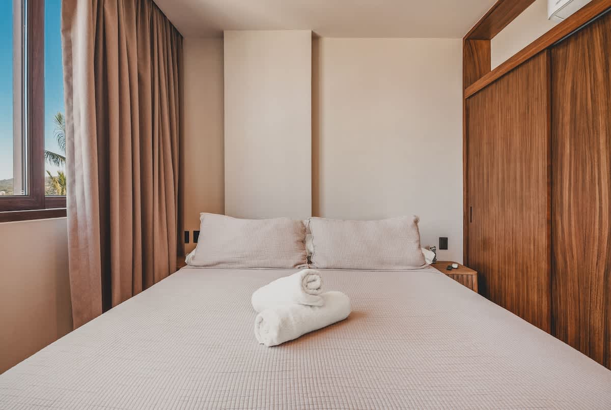 View of the bedroom from the foot of the bed, showing a queen-sized bed with light bedding, two rolled white towels, a large window with light brown curtains, and a wooden built-in wardrobe on the right.