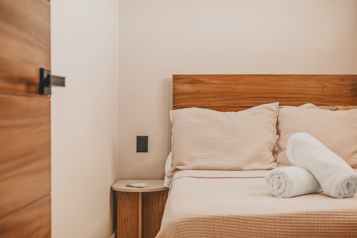 Cozy bedroom featuring a wooden headboard, light beige bedding, and two white rolled towels on the bed, with a simple wooden nightstand and a dark wall outlet beside it.