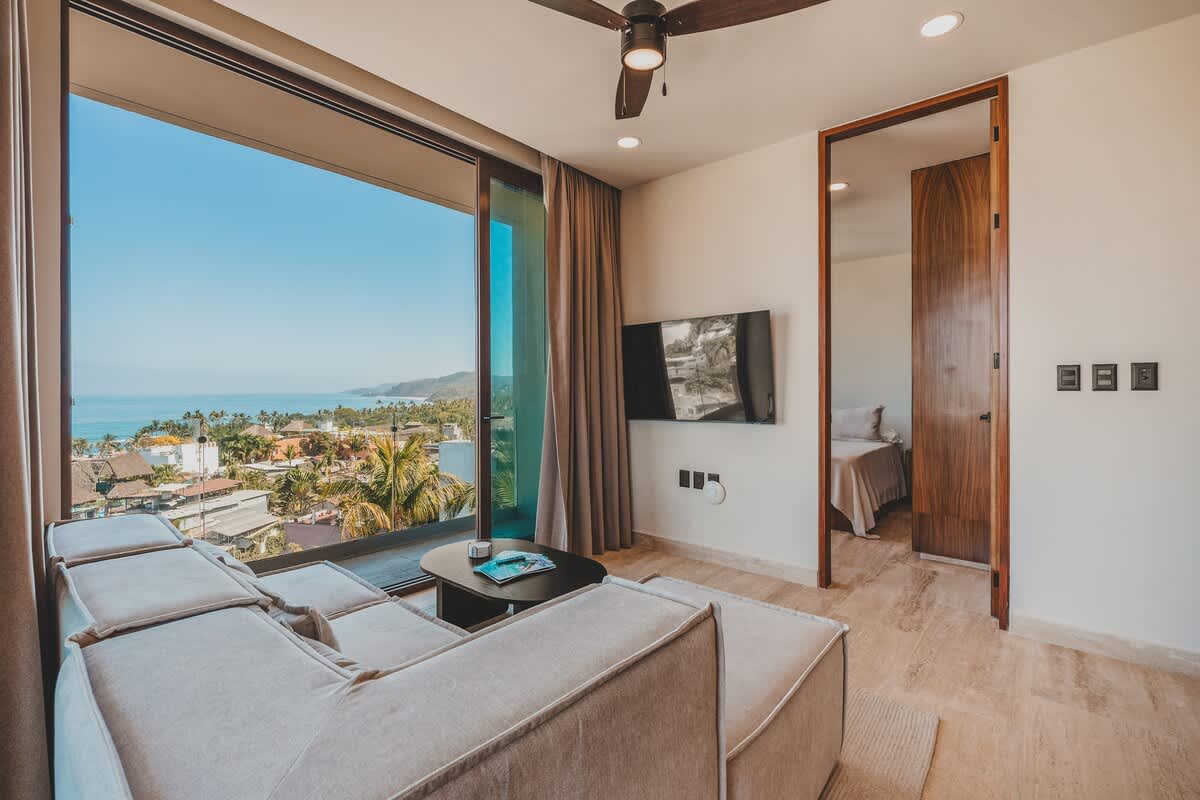 Modern living room with a light beige sectional sofa, a wall-mounted flat-screen TV, and a dark ceiling fan, with a wooden doorway leading to a bedroom and large windows offering ocean views.