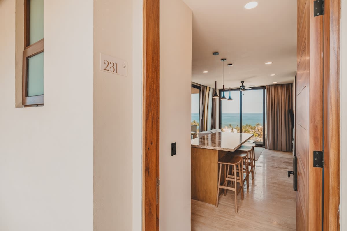 A bright entryway with room number '231' on the wall, opening into a spacious living area featuring a large kitchen island with bar stools. Floor-to-ceiling windows offer expansive ocean views, and the flooring is light-colored tile.