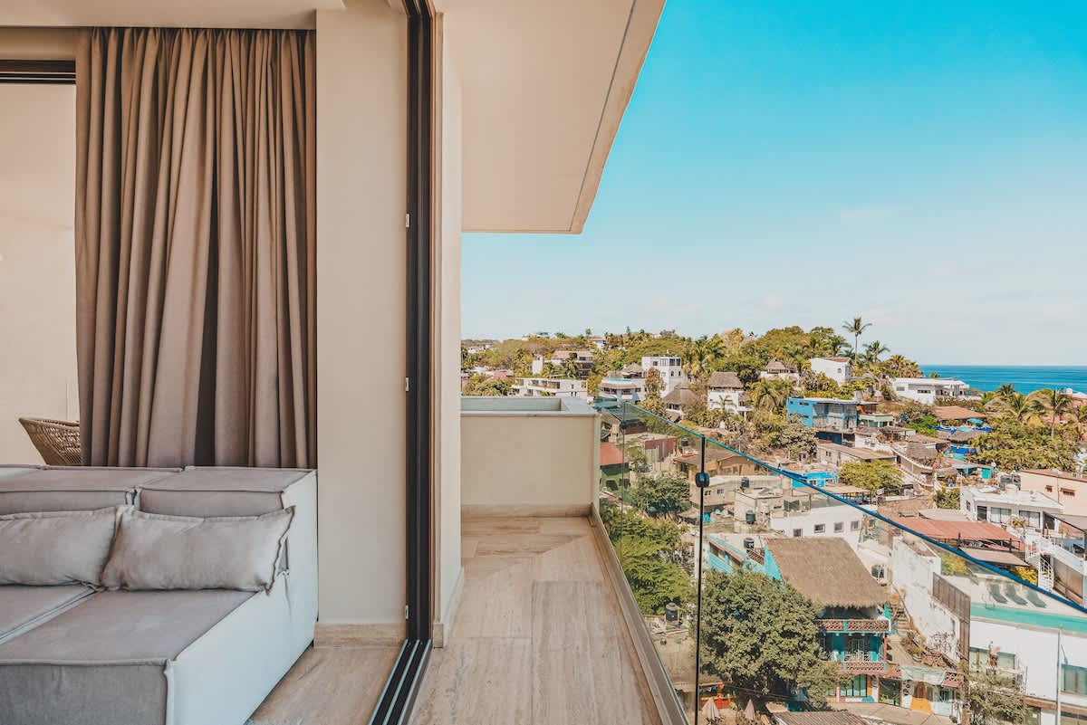 A spacious balcony with a clear glass railing offers a sweeping view of a bustling coastal town and the distant ocean under a bright blue sky.